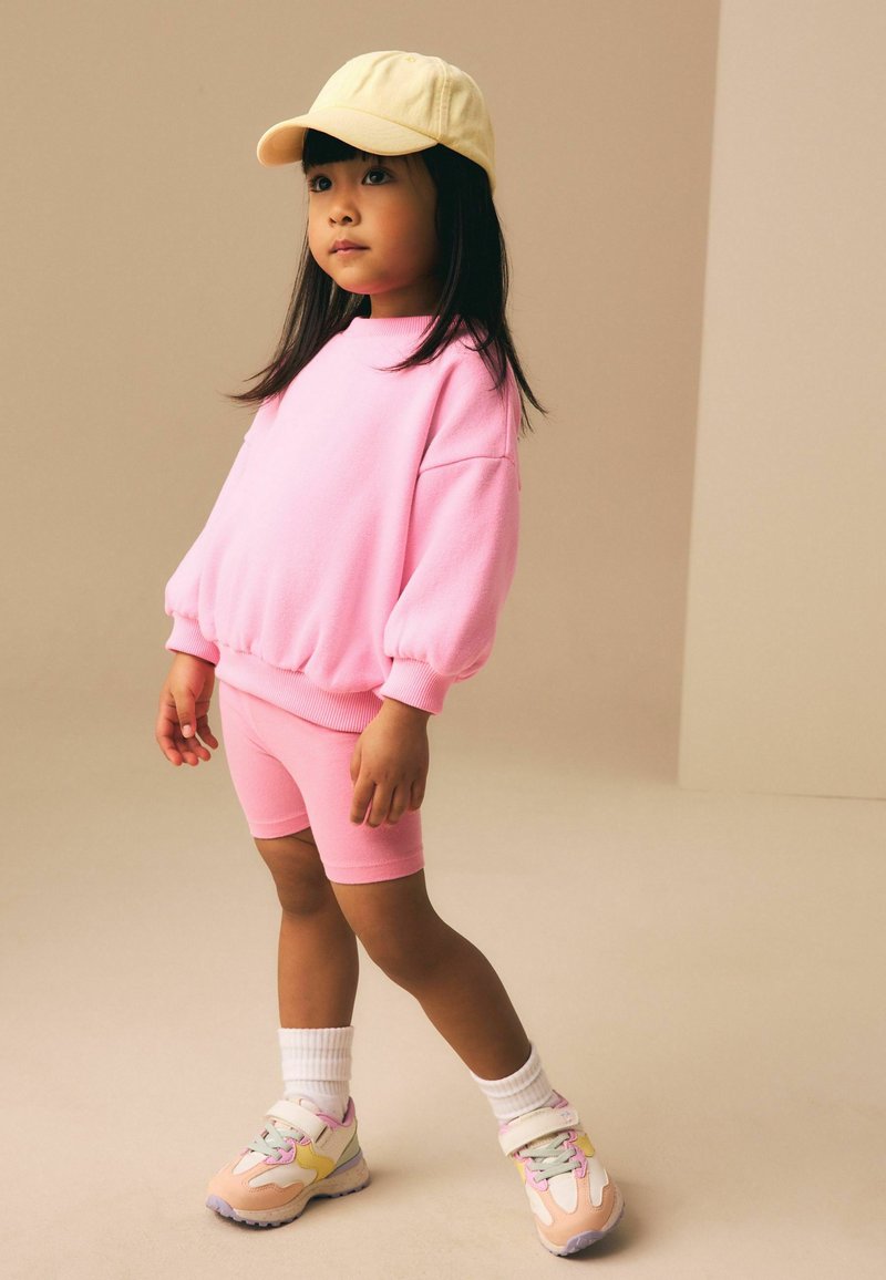 Junges Mädchen mit gelber Mütze, rosa übergroßem Sweatshirt, passenden Shorts, weißen Socken und pastellfarbenen Sneakers, steht vor einem einfarbigen Hintergrund.