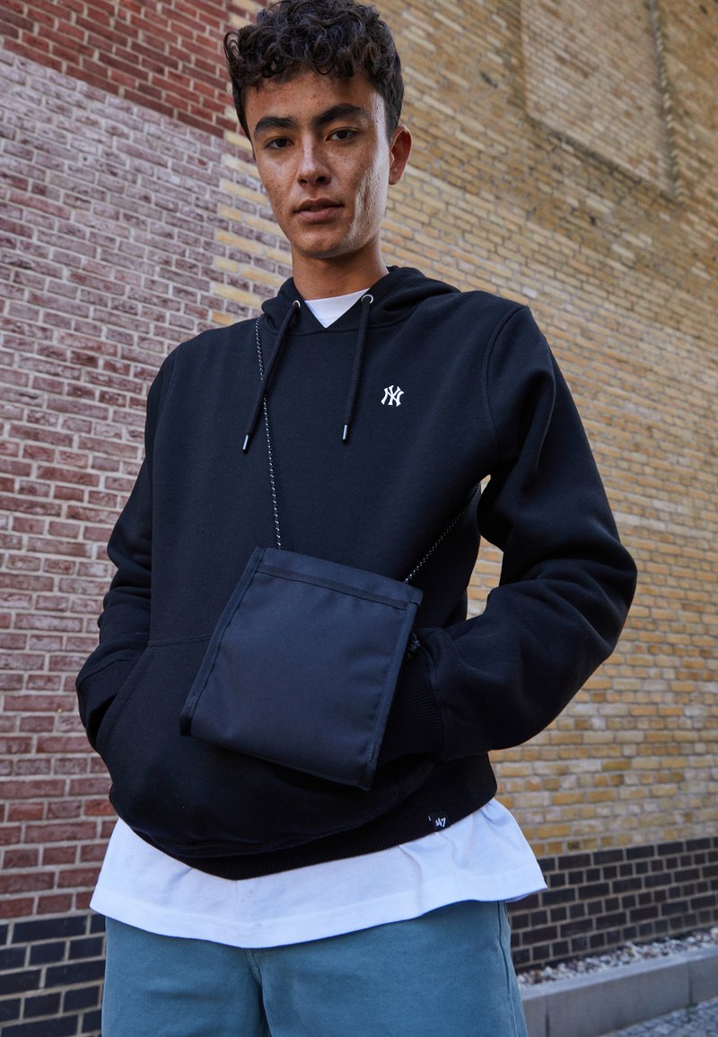 Zwarte hoodie met een voorzak en een capuchon met trekkoorden, gedragen over een wit shirt. Heeft een klein logo op de borst en een afneembare zwarte pouch.