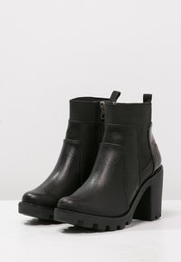 Bottes noires à la cheville en matériau synthétique, dotées d'un talon chunky haut, d'une fermeture éclair sur le côté et d'un détail texturé sur le dessus.