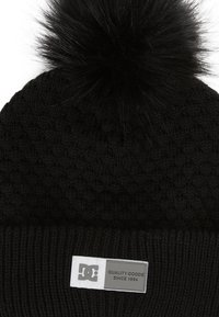 Black knitted beanie with a textured pattern, topped with a black faux fur pom-pom. Features a rectangular logo patch at the front.