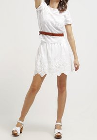 Weißes Stricktop mit Spitzenakzenten, kombiniert mit einem weißen Rock mit besticktem, gewelltem Saum. Gewebter brauner Gürtel und weiße Plateau-Sandalen.