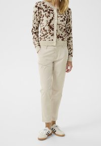 Cardigan beige avec des taches marron, fermeture à boutons, associé à un pantalon beige clair et des baskets blanches avec des rayures noires.