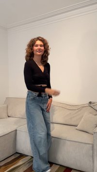 Top preto de mangas longas envolvente com decote em V, combinado com jeans de perna larga azul claro. O fundo inclui um sofá texturizado em bege.
