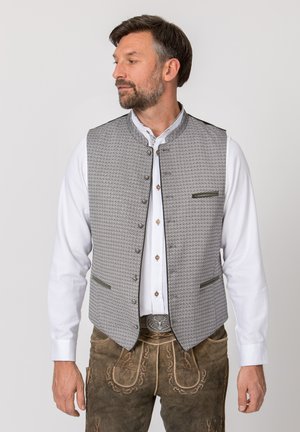 Mann trägt traditionelle bayerische Tracht mit bestickter brauner Lederhose, weißem Hemd und gemusterter grauer Weste, schaut zur Seite.