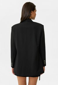 Zwarte op maat gemaakte blazer met gestructureerde schouders, lange mouwen en knoopaccenten bij de manchetten. Glad materiaal en vloeiende silhouet.