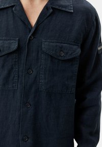 Camisa de botón azul oscuro con cuello, dos bolsillos en el pecho con solapas y mangas largas. Fabricada en una tela texturizada con detalles sutiles.