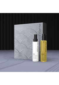 Due bottiglie di skincare, una bianca e una gialla, sono posizionate di fronte a una scatola grigia testurizzata con motivi a onda su una superficie liscia.
