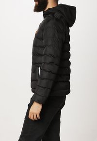 Chaqueta acolchada negra con capucha, con un diseño de acolchado y un parche con el logo en el pecho. La tela parece suave y ligera.