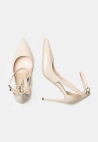 Décolleté in vernice beige con punta affusolata, cinturino alla caviglia, design a ritaglio e tacco a stiletto sottile, dotato di una fibbia decorativa.