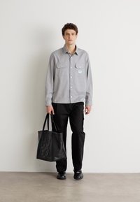 Gray button-up shirt with two chest pockets, paired with black pants and black shoes, holding a black tote bag with embossed logo.