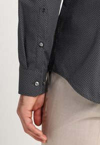 Svart skjorta med vita prickar och knappärmar, visad på en delvis synlig hand. Slät tyg med kontrasterande vita prickar.