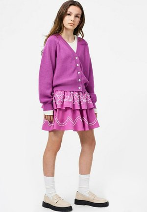 Giovane donna che indossa un cardigan viola con bottoni e una gonna a strati a fantasia abbinata, con calze bianche e scarpe beige, in piedi su uno sfondo bianco.