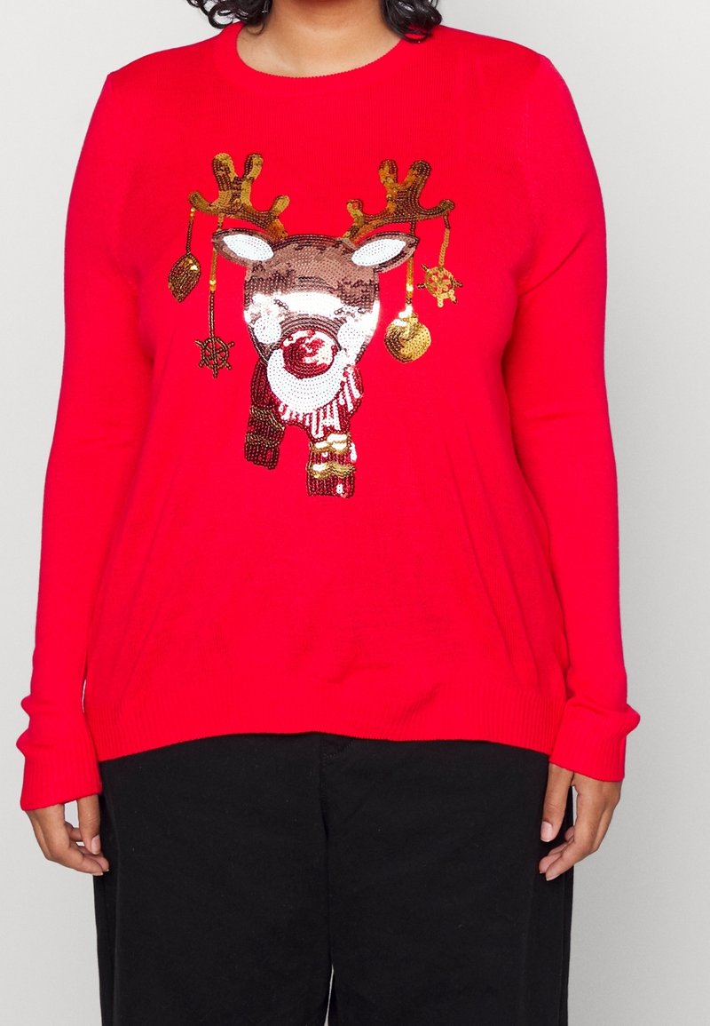 Pull rouge avec un visage de renne à paillettes portant une écharpe et des bois décorés, porté par une personne avec un pantalon noir.