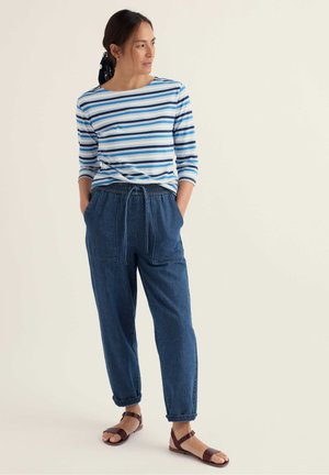 Mujer con camisa de rayas azules y blancas de manga larga, pantalones vaqueros holgados y sandalias marrones, de pie con las manos en los bolsillos, mirando hacia la izquierda.