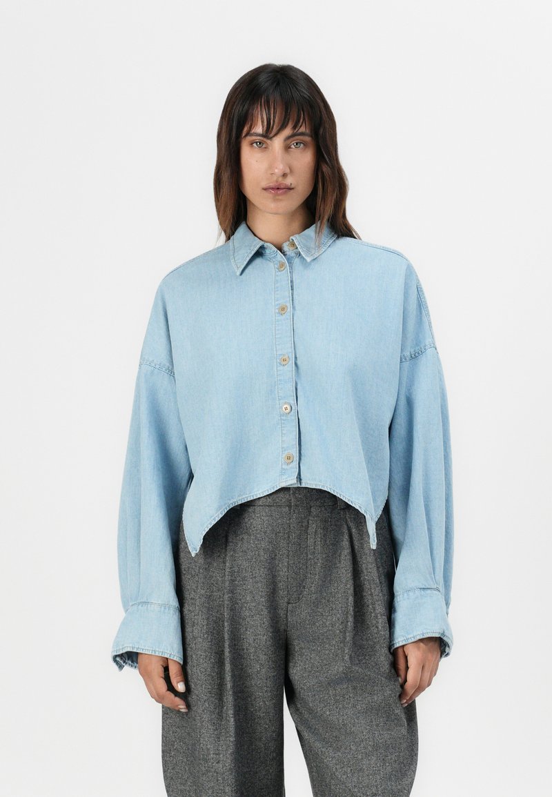 Chemise en denim bleu clair, courte avec une fermeture à boutons sur le devant, un col pointu et des manches longues. Présente un ourlet arrondi et une coupe décontractée.