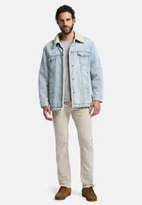 Ljusblå denimjacka med en beige schäferkrage, två bröstfickor och en avslappnad passform, kombinerad med beige byxor och bruna skor.