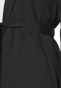 Manteau noir enveloppant avec une texture lisse, une ceinture intégrée et des poches latérales, offrant une silhouette épurée et cintrée.