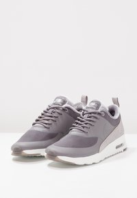 Nike-sneakers i ljusgrått, med en överdel i mesh, swede-accenter, texturerade överlägg och en vit dämpad sula med en synlig luftenhet.