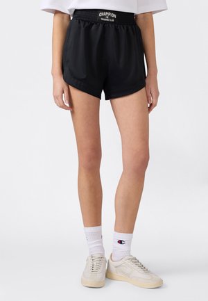 Osoba ubrana w czarne spodenki Champion Training Club, białe skarpetki Champion i kremowe tenisówki, stojąca na jednolitym tle.