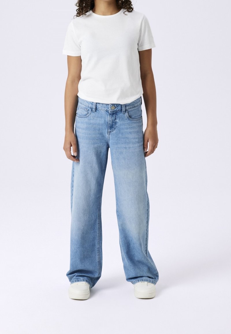 Jeune personne portant un t-shirt blanc uni à manches courtes, un jean ample bleu clair et des baskets blanches, debout devant un fond uni.