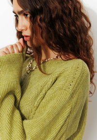 Pull en tricot vert avec un motif texturé, manches larges et poignets côtelés. Collier en chaîne dorée visible à proximité.