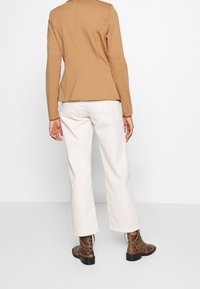 Personne portant une veste beige, un pantalon blanc à jambes larges et des bottines à motif peau de serpent marron, debout devant un fond blanc uni.