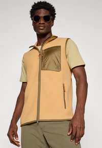 Beige fleece vest met een olijfgroene zak en ritsdetails. Draagt over een lichtgroen shirt met korte mouwen. Textuurstof.