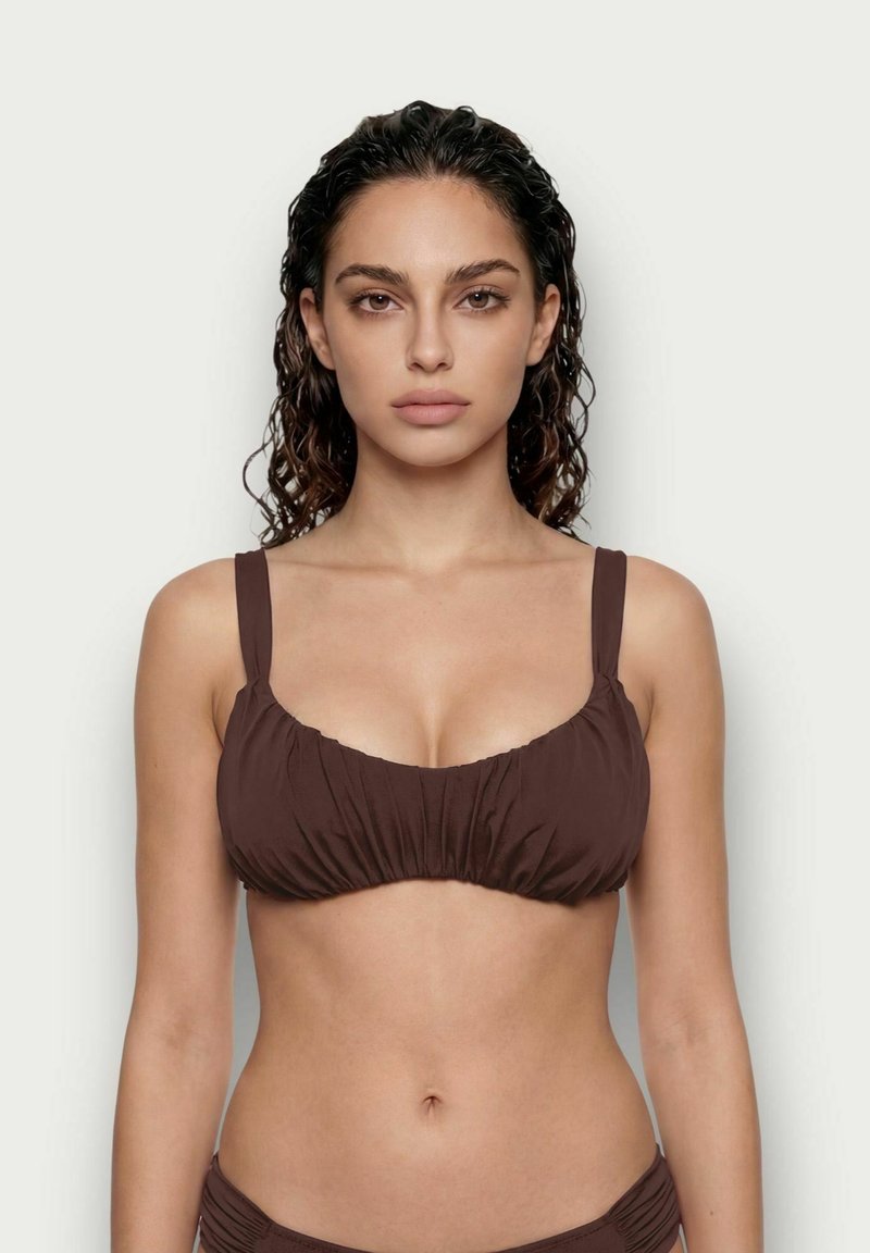 Mujer con el pelo mojado que lleva un top de bikini marrón, mirando hacia adelante sobre un fondo liso y claro.
