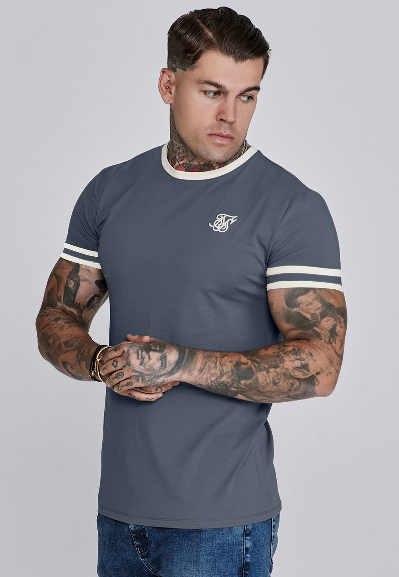 Uomo con tatuaggi su braccia e collo che indossa una maglietta grigia con bordi bianchi e loghi, con le mani incrociate, guarda a sinistra, in piedi contro uno sfondo neutro.