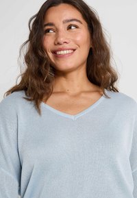 Femme souriante avec des cheveux bruns bouclés portant un pull V bleu clair, regardant légèrement sur sa gauche devant un fond uni.