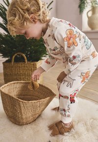 Children's pajama set in cream with colorful gingerbread and festive patterns; textured cotton fabric, matching soft slippers, and woven basket nearby.