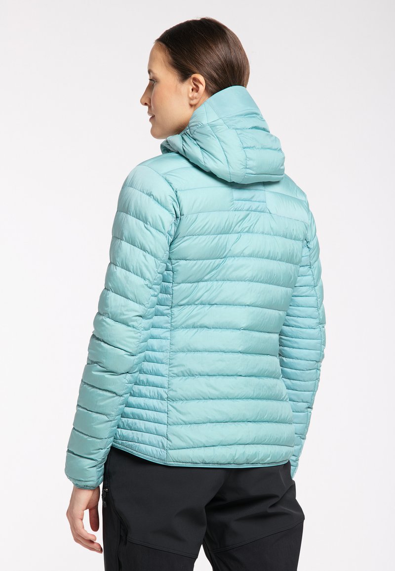 Mujer con una chaqueta acolchada con capucha de color azul claro, vista desde la espalda, combinada con pantalones negros, contra un fondo blanco liso.