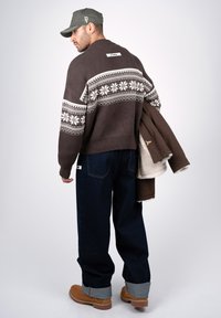 Homme portant un pull sombre à motifs, un jean foncé avec des revers, des bottes couleur tabac, une casquette verte, tenant une veste en shearling marron sur l'épaule.