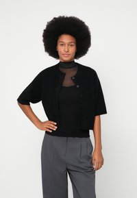 Black cropped cardigan made of soft fabric with short sleeves and button closure, worn over a black top and paired with gray trousers.