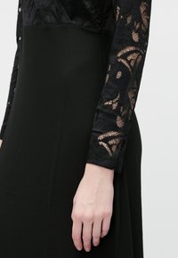 Svart kjole med lange ermer og blondedetaljer samt en glatt, ensfarget skjørt. Den teksturerte blonden kontrasterer med det matte stoffet.