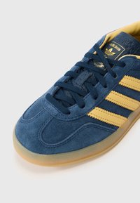 Zils zamšādas sports apavu ar dzeltenām svītrainām detaļām, gumijas zolīti un biezām auklām. Aprīkots ar teksturētu augšdaļu un logotipu mēlē.