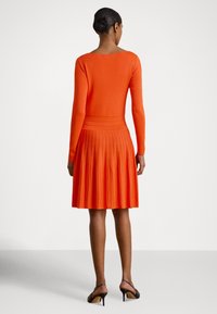 Moteris, stovinti nugara į kamerą, dėvi ilgomis rankovėmis oranžinę, klostuotą suknelę ir juodus basutus ant pakulnių, baltame fone.