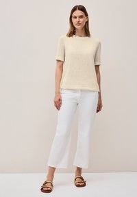 Beige texturovaný svetr s krátkým rukávem, kombinovaný s bílými širokými kalhotami a hnědými sandály na pásky, nošený na neutrálním pozadí.