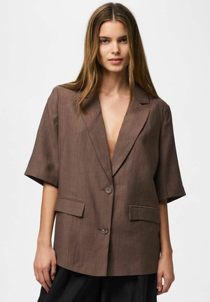 Jeune femme aux longs cheveux bruns portant un blazer marron ample à manches courtes et un pantalon noir sur un fond uni.