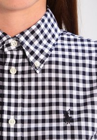Camicia a scacchi bianca e nera con colletto button-down, bottoni in plastica e logo ricamato sul petto.