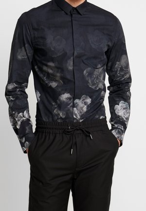 Mann, der ein schwarzes Hemd mit Knöpfen und dunkelgrauem Blumenmuster sowie schwarze Hose mit Kordelzug trägt, Hände in den Taschen, vor schlichtem Hintergrund.