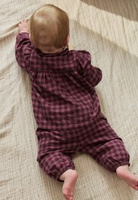 Combinaison courte à carreaux bordeaux et rose foncé avec manches longues et poignets élastiques, vue de dos sur une surface en tissu beige texturé.