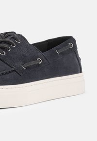 Καθημερινό παπούτσι τύπου boat από μπλε σουέντ ναυτικού χρώματος με λευκή λαστιχένια σόλα, μαύρα κορδόνια και μεταλλικές τρύπες στα πλαϊνά πάνελ.
