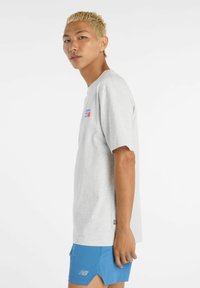 Grå kortärmad t-shirt med rund halsringning, prydd med en färgglad logotyp på bröstet. Bärs tillsammans med blå shorts. Slät textur.