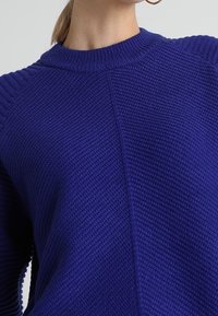 Pull-over en tricot bleu royal vif avec un motif texturé, un col côtelé et des manches raglan. Vue en gros plan mettant l'accent sur les détails du matériau.