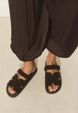 Feet wearing dark brown woven sandals with straps, paired with loose, dark brown pants on a light floor.