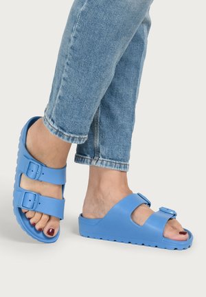 Feet wearing blue open-toe slide sandals with adjustable buckles, paired with cropped blue denim jeans and red painted toenails.