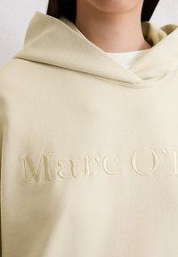 Kreemivärvi kapuutsiga särk, millel on pehme tekstuur, esiküljel tikandiga logo ja mugav, ülekaaluline lõige.