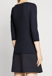 Frau in einem navyblauen, gerippten Langarmoberteil und lockeren dunklen Shorts, die mit dem Rücken zur Kamera vor einem einfarbigen Hintergrund steht.