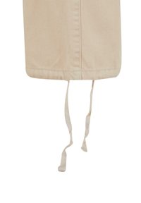 Beige stoffforkle med en teksturert overflate, med en kant og to lange, tynne bånd som strekker seg fra bunnen for festing.